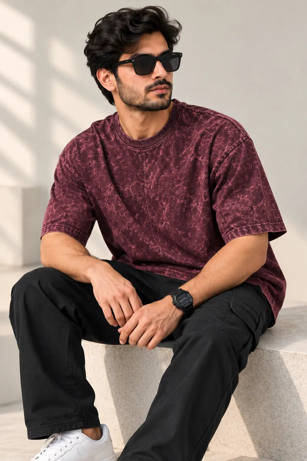 Man seated wearing a maroon acid wash oversized t-shirt with black cargos and white sneakers