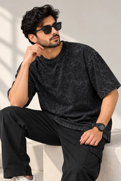 Man seated wearing a black acid wash oversized t-shirt with black cargos, sunglasses and white sneakers